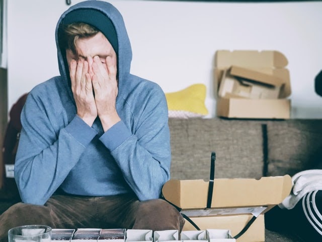 A person in a blue hoodie sitting on a couch with their head in their hands.