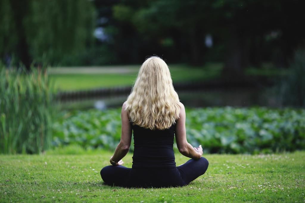 A woman with long blonde hair sitting in the grass near a pond practicing meditation, facing away from the camera.