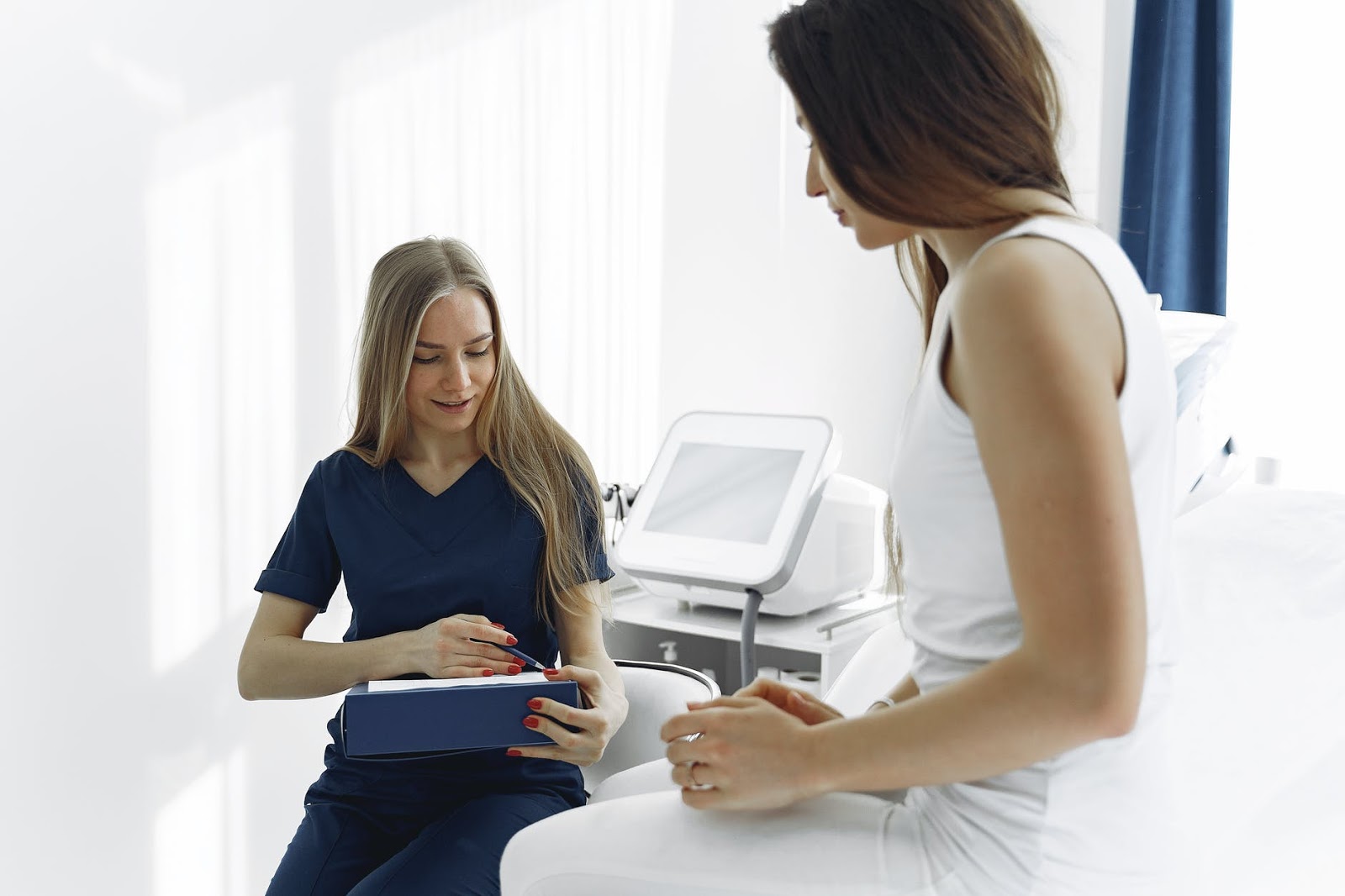 A nurse wearing blue scrubs holds a binder and talks with a patient, who is sitting down on an exam table.