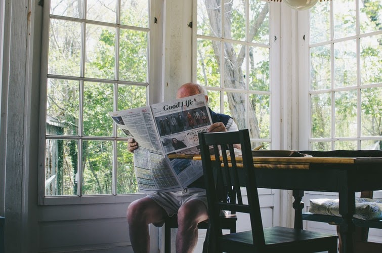 A senior citizen reads the newspaper in their kitchen.