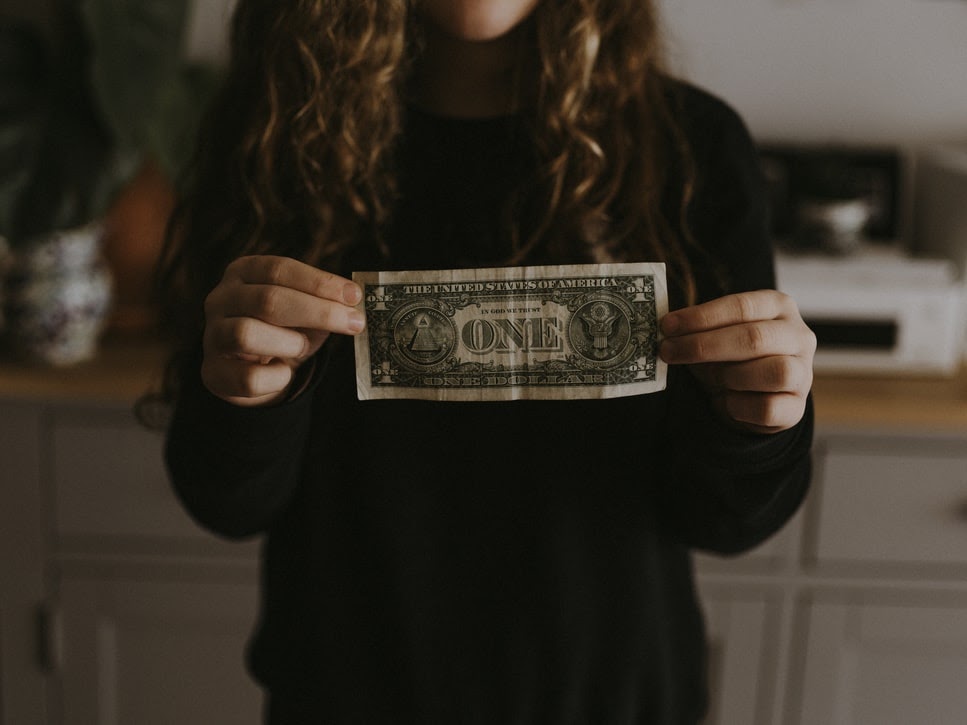 A person with long curly brown hair wears a black turtleneck sweater and holds a single American dollar bill in their hands.